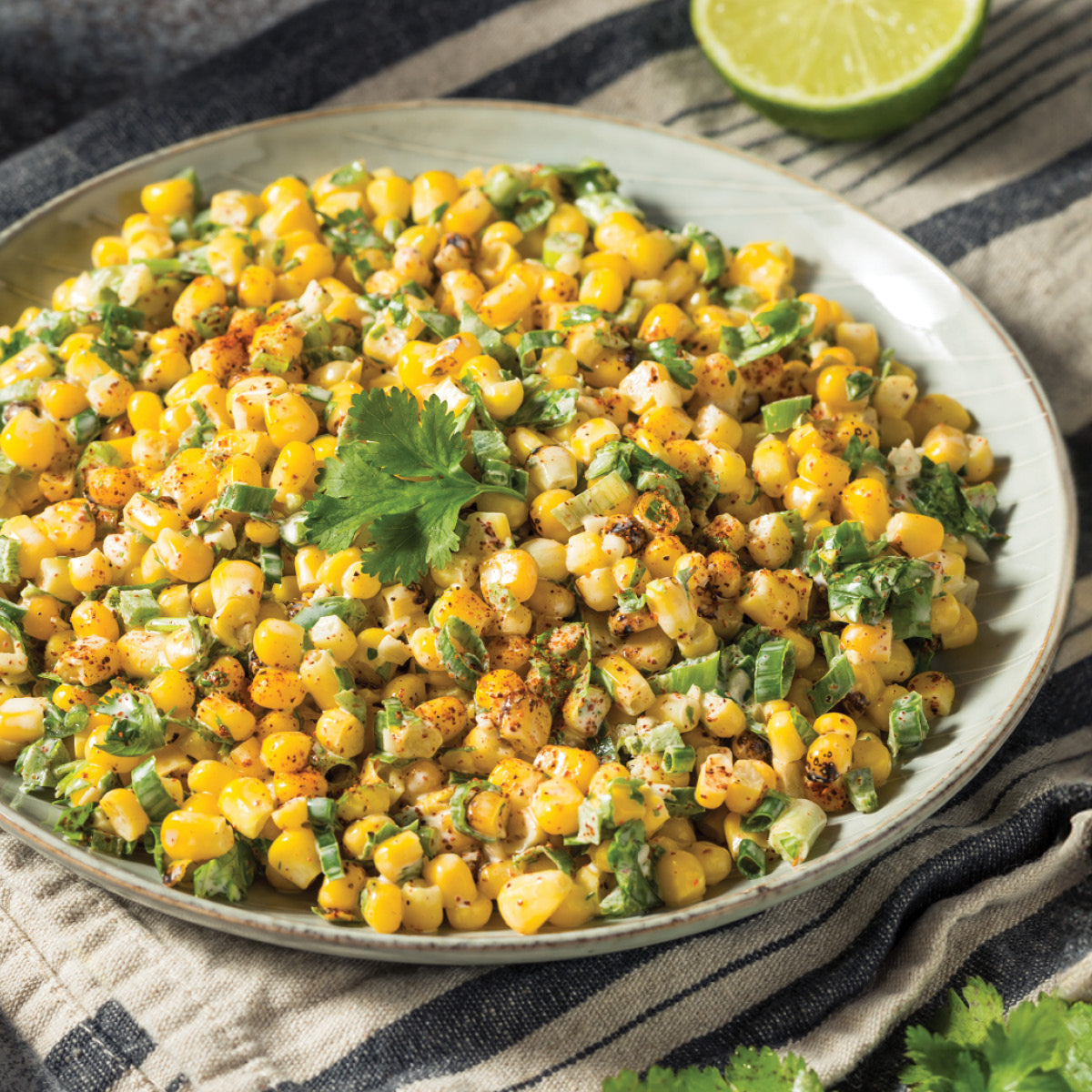 A plate of Mexican street corn salad garnished with cilantro, featuring roasted corn, chopped green onions, and spices, with a lime half in the background on a striped cloth.